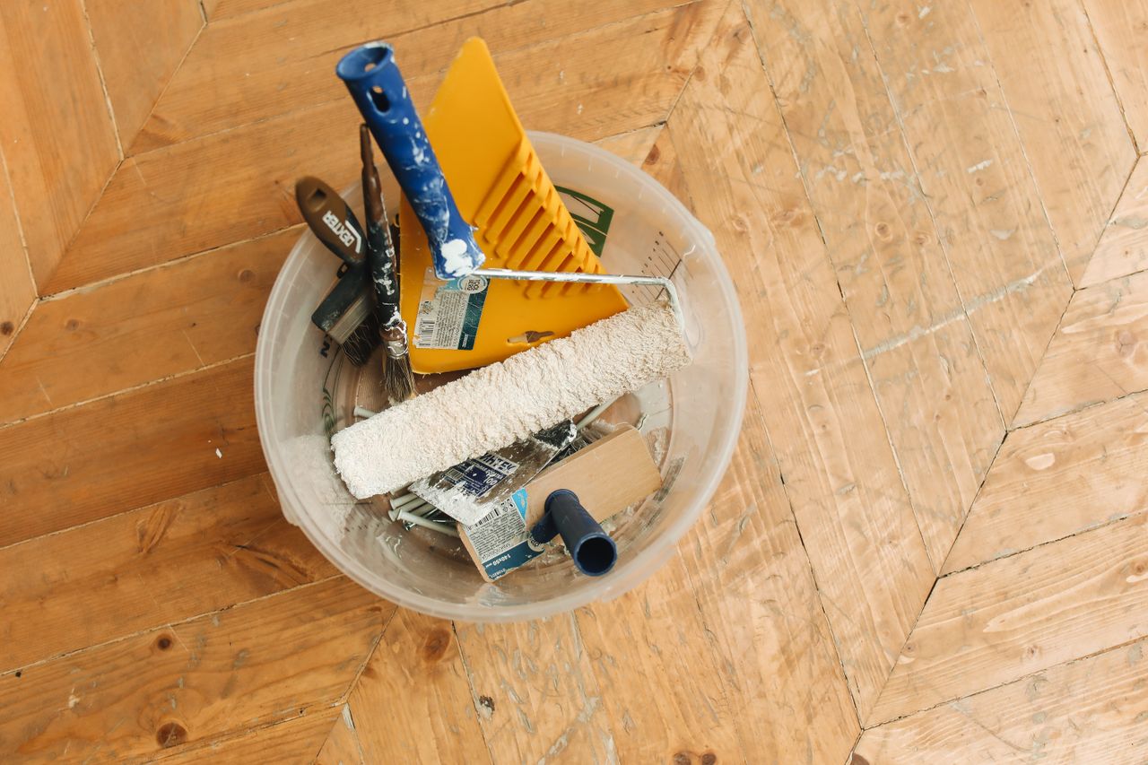 Strona główna A collection of home renovation tools in a bucket on a wooden floor, ready for a painting project.